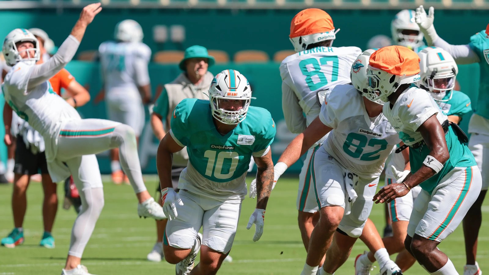 Miami Dolphins players running a drill in helmets and practice jerseys during a training camp session, with coaching staff and equipment visible along the sideline.