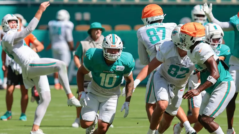 Miami Dolphins players running a drill in helmets and practice jerseys during a training camp session, with coaching staff and equipment visible along the sideline.